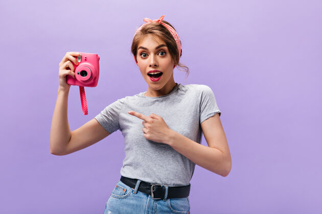 紫色穿着灰色衬衫的惊讶女人拿着粉红色的相机可爱的年轻女孩留着时髦的发型 戴着头带摆姿势孤立的背景头发成人休闲