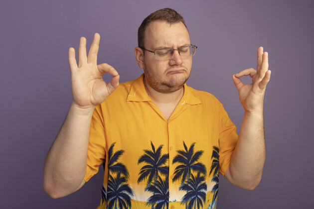 站着一个戴着眼镜的男人穿着橘色衬衫 闭着眼睛站在紫色的墙上做着冥想的手势戴男人闭上