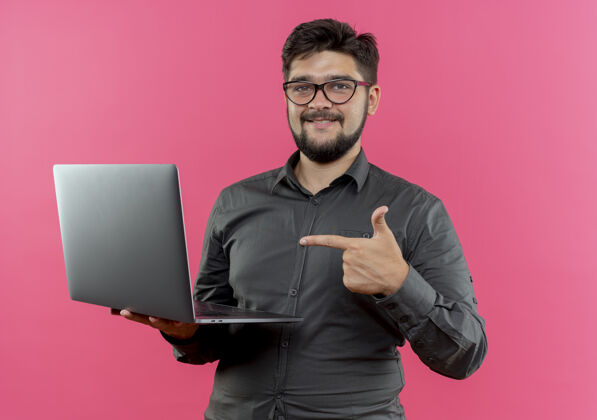 商务人士高兴的年轻商人戴着眼镜拿着笔记本电脑指着隔离在粉红色背景上戴年轻人笔记本电脑