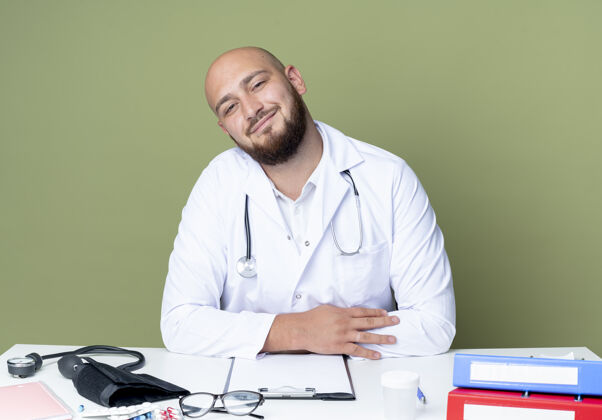 医疗高兴的年轻秃头男医生穿着医用长袍和听诊器坐在办公桌前工作 医疗工具隔离在绿色背景上办公桌秃头医生