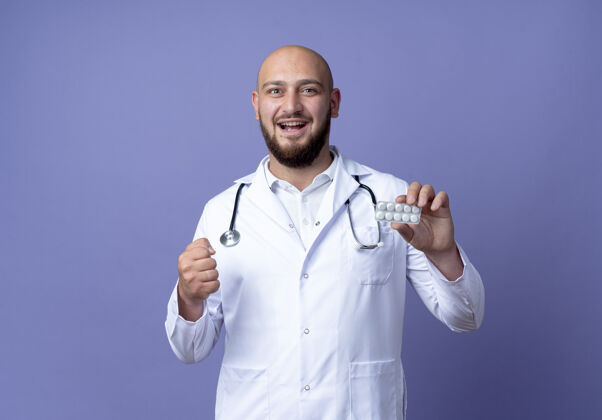 年轻快乐的年轻秃头男医生穿着医用长袍和听诊器拿着药片 在蓝色背景上显示“是”的手势高兴穿医疗