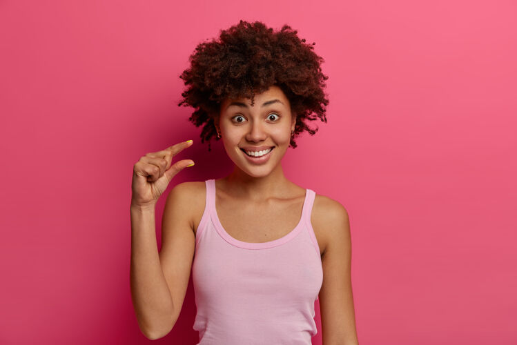 形状积极满足的黑皮肤年轻女子做小尺寸的标志 展示很少的东西 手势和形状的对象 穿着休闲背心 隔离在粉红色的墙壁上黑发小室内