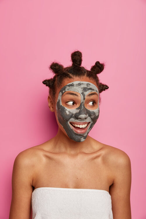 光滑开朗的非裔美国女人的垂直镜头有着清新干净的皮肤 看着旁边 涂上粘土面膜 享受面部护理 站在浴巾里 隔离在粉红色的墙上美容养颜年轻化请应用