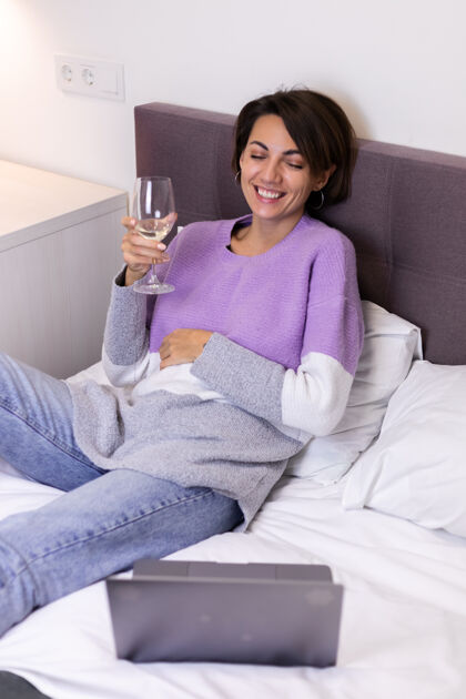 房子快乐的女人穿着暖和的套头衫 躺在床上 一个人喝着一杯葡萄酒 休息着看喜剧电影 笑着笑着笔记本女孩嘴