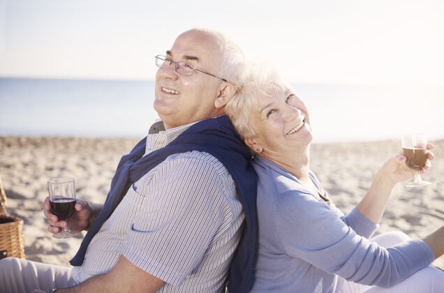 无忧无虑老年人背靠背坐着喝酒老年夫妇在沙滩上 退休和暑假的概念照明海滩婚礼