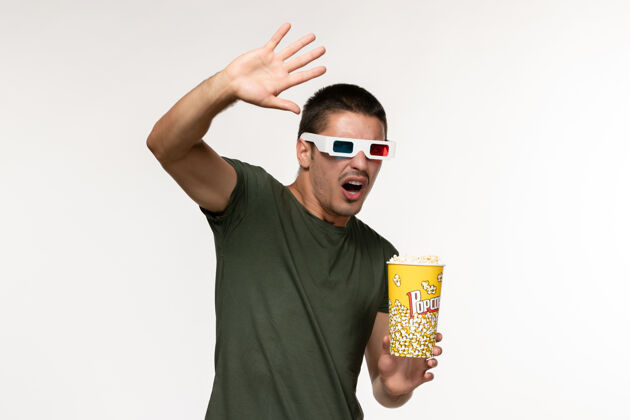 观看正面图身穿绿色t恤的年轻男子戴着d型太阳镜拿着爆米花在白墙上看电影孤独电影院男电影男人电影观点