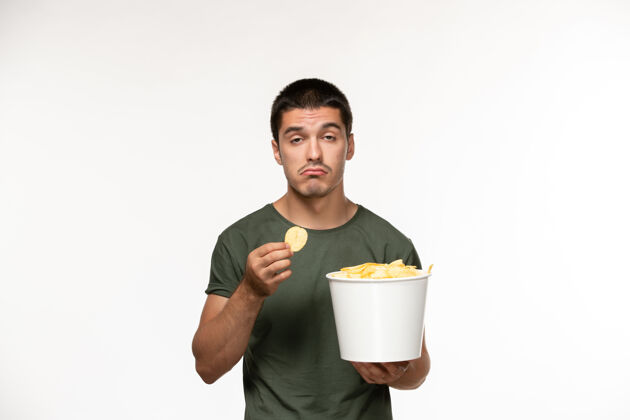 成人正面图：穿着绿色t恤的年轻男性 带着土豆cips 在白墙上吃饭 孤独的人在电影院电影电影服务员