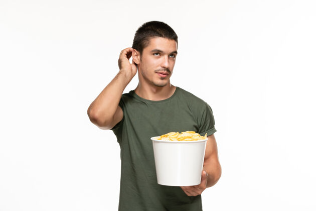 微笑正面图身穿绿色t恤的年轻男性手持土豆cips 在白墙上看电影孤独的电影成人土豆视图