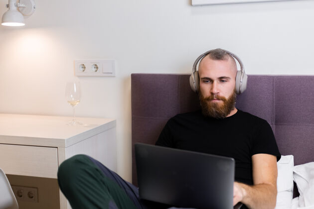 手机一个留着大胡子的白种人戴着耳机在卧室的床上工作 在家里用笔记本电脑 打字 思考放松胡须网络