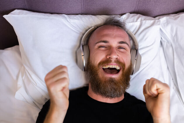 年轻快乐的白人男子戴着耳机躺在床上听他最喜欢的音乐 独自休息 跳舞享受家伙休息
