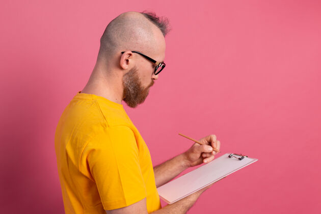 成人年轻的留着胡须的员工男士休闲装在粉色背景下做笔记文件男空