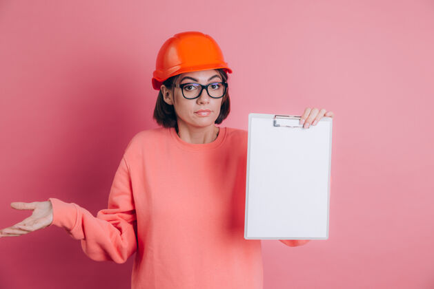 工作女建筑工人拿着白色的空白标牌 背景是粉色的戴着建筑头盔女人举起双手耸耸肩 毫无头绪建筑女士帽子