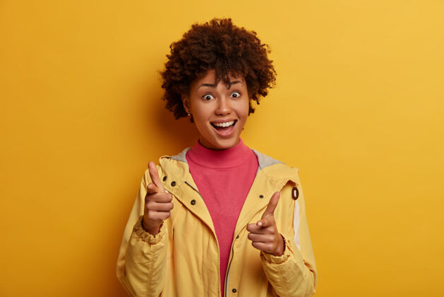 感觉嘿 你 注意客户选择概念积极的非洲裔美国女孩用快乐的表情点 选东西 选人 穿休闲服 隔离在黄色的墙上单色休闲选择