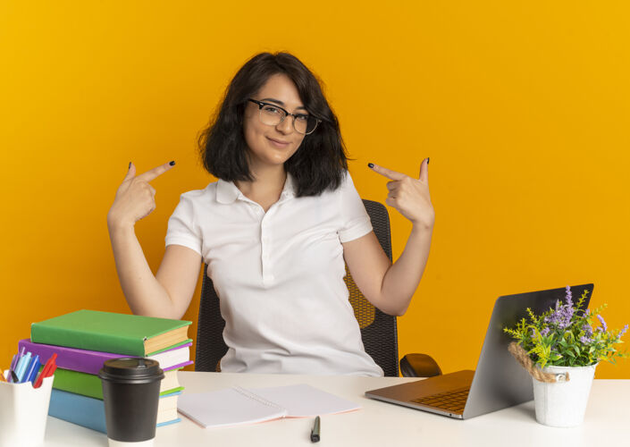 复制年轻漂亮的高加索女生戴着眼镜坐在书桌旁 手里拿着学习工具 指着橙色的自己 留着复印空间请穿点