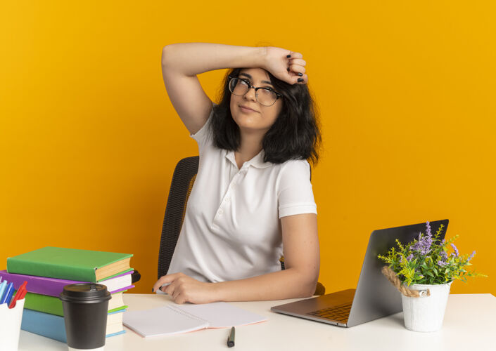 空间年轻漂亮的高加索女生戴着眼镜坐在书桌旁用学习工具把胳膊放在额头上看着橙色的侧面和复印空间年轻手臂额头