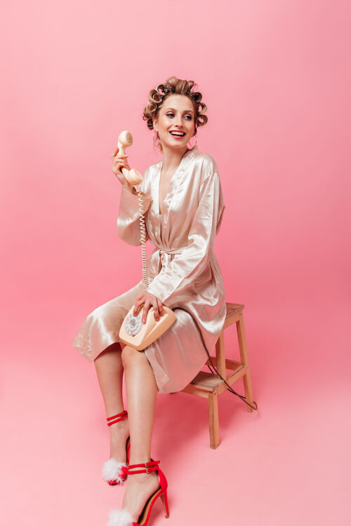 时尚穿着粉色长袍的迷人女人微笑着 坐在椅子上 在隔离的墙上拿着电话成人衣服沟通