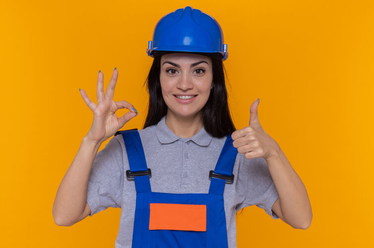 积极身穿施工制服 头戴安全帽的年轻建筑工人高兴而积极地向上竖起大拇指 橙色的墙上立着“ok”标志女人快乐年轻