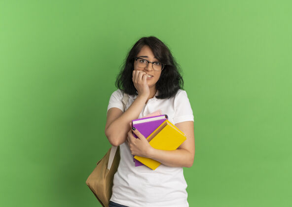 女生年轻焦急漂亮的高加索女学生戴着眼镜背着书包咬着手指头拿着书放在绿色的有复印空间的地方焦虑复制漂亮