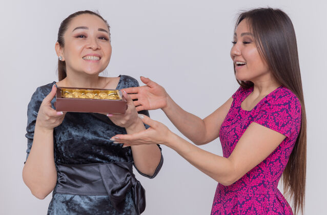 兴奋两位快乐而兴奋的亚洲妇女手持一盒巧克力糖果 站在白色的墙上 喜气洋洋地微笑着庆祝国际妇女节国际庆祝巧克力