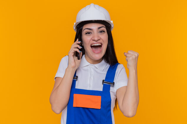 建设者身着施工制服 头戴安全帽的年轻建筑工人站在橙色的墙上 一边用手机聊天 一边高兴地握紧拳头握紧拳头电话