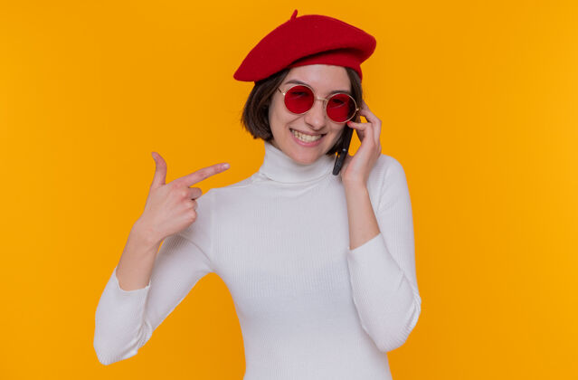 年轻快乐而积极的年轻女子 短发 白色高领毛衣 头戴贝雷帽 戴着红色太阳镜 用食指指着站在橙色墙上的手机贝雷帽目录太阳镜