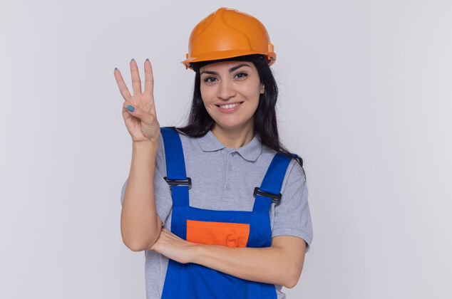头盔身穿建筑制服 头戴安全帽的年轻建筑妇女微笑着看着前面 手指站在白色的墙上 展示着三号人物表演建筑建设者