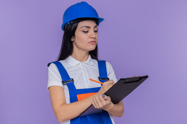 拿着身穿建筑制服 头戴安全帽的年轻建筑工人站在紫色的墙上 手里拿着剪贴板 写着一些看起来很自信的东西写作建筑女人