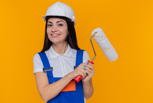 抱身着施工制服 戴着安全帽 拿着油漆辊 面带微笑 自信地站在橙色墙壁上的快乐的年轻建筑妇女头盔快乐建筑
