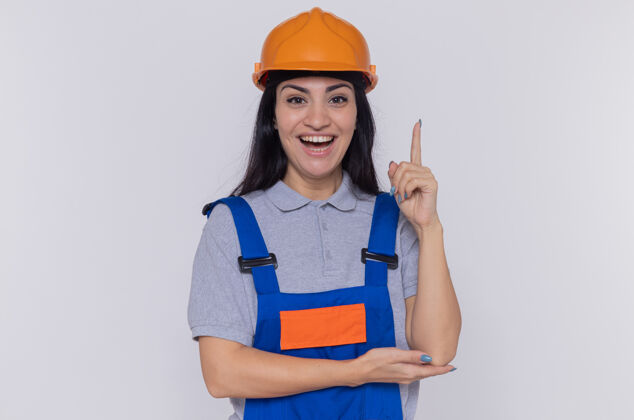 年轻身穿建筑制服 头戴安全帽的年轻建筑工人面带微笑地看着前面 食指站在白色的墙上目录女人建设者