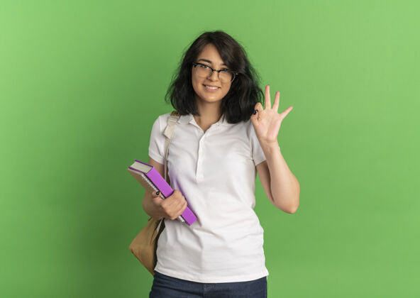 标志年轻笑容可掬的白人女学生戴着眼镜 背着书包 手势ok 手语拿着书放在绿色的纸上 留着复印空间包空间女生