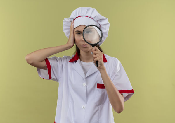 持有身着厨师制服的年轻自信的白人厨师女孩手持放大镜 将手放在有复印空间的绿地上手放大镜头
