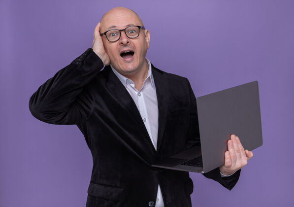 迷糊穿西装戴眼镜的中年秃头男子拿着笔记本电脑一边看一边困惑地用手捂着头 误站在紫色的墙上笔记本电脑拿着头