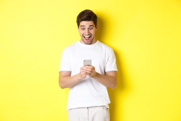 成年人惊讶的男人在手机上阅读短信 看着智能手机屏幕 惊讶而快乐男人时尚黄色