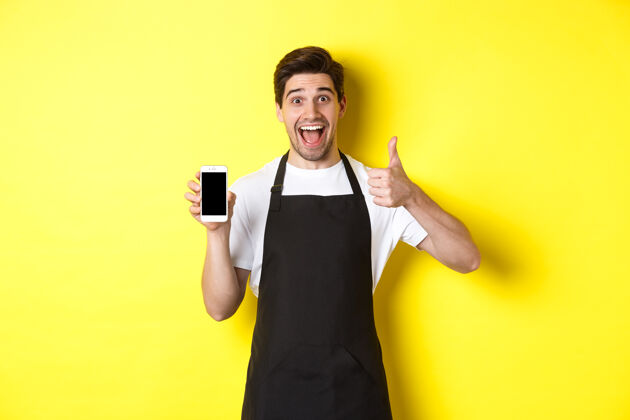 展台身着黑色围裙的快乐咖啡师展示智能手机屏幕 竖起大拇指 推荐咖啡馆应用程序 站在黄色背景上成人黄色年轻