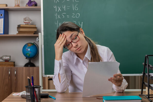 工作戴着眼镜的年轻女教师坐在课桌旁 拿着空白的书页 在教室的黑板前检查作业 看上去很累 而且工作过度教室空白过度工作