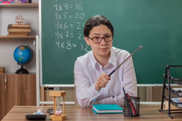 女人戴着眼镜的年轻女老师面带严肃的表情 指着坐在教室黑板前的课桌上讲解课文的东西老师指黑板
