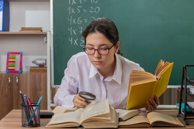 黑板戴眼镜的年轻女教师坐在教室黑板前的课桌旁 用放大镜看书 感到困惑和不快桌子教室不高兴