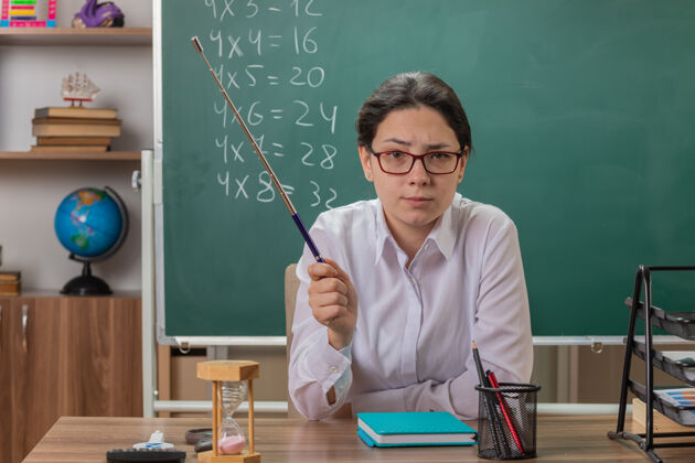 黑板年轻的女老师戴着眼镜 神情严肃自信地看着前面 手里拿着指针 坐在教室黑板前的课桌上讲解课文穿解释前面