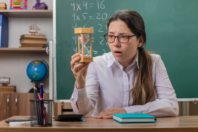 课年轻的女老师戴着沙漏眼镜看着它 惊讶地坐在教室黑板前的课桌上备课沙漏老师年轻人