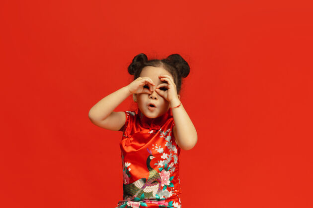 旗袍摆出可爱的姿势 寻找礼物…亚洲可爱的小女孩在传统服装隔离在红墙庆祝 人类的情感 节日的概念复制空间问候情感年轻