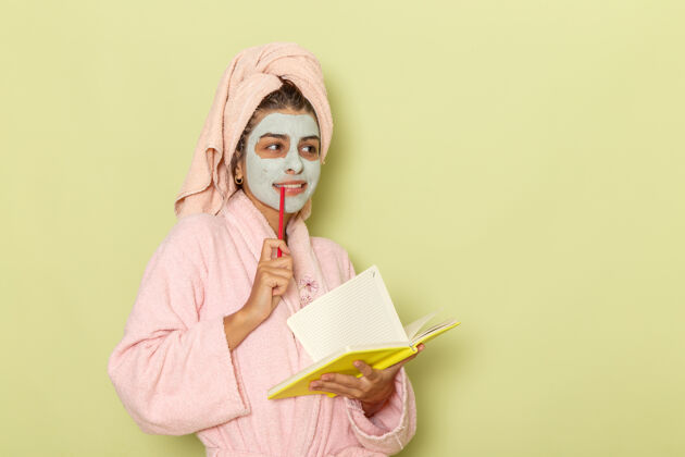 面具正面图穿着粉红色浴袍的年轻女性在绿色的表面上写下笔记和思考美丽自我照顾浴袍