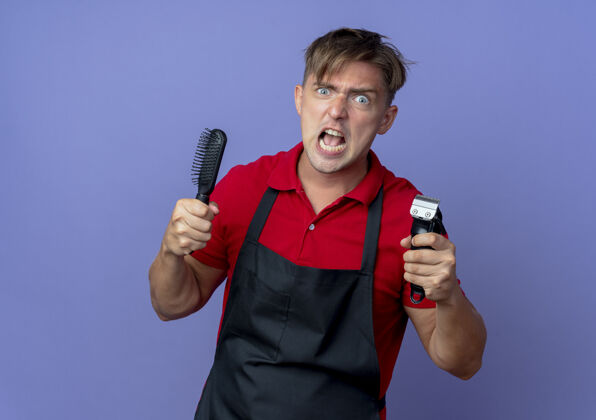 年轻年轻狂暴的金发男理发师穿着制服 拿着梳子和理发器孤立地放在紫色的空间和复制空间愤怒持有剪子