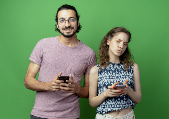 不高兴一对年轻漂亮的夫妇 一男一女 微笑着拿着智能手机站在他不高兴的女友旁边 手机越过绿墙女人年轻抱着