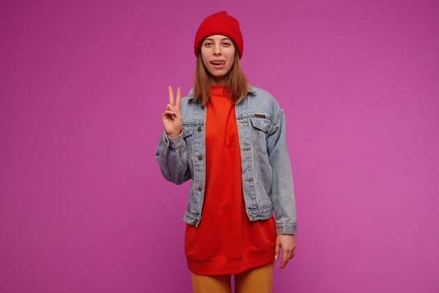单独有魅力的女人的肖像 深色长发穿着牛仔裤夹克 黄色裤子 红色毛衣和帽子在紫色的墙上显示和平标志和舌头和平完美女孩