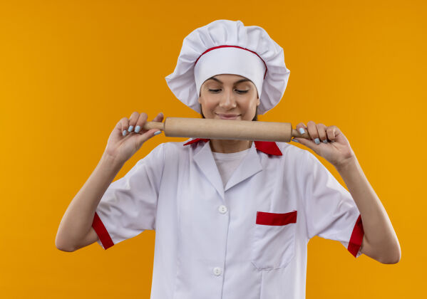 别针身着厨师制服的年轻高加索厨师女孩高兴地拿着擀面杖 看着被隔离在橙色空间里的擀面杖请卷复制