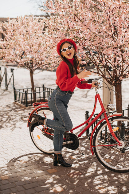 女性年轻时尚的女孩在樱花旁和红色自行车合影穿着羊毛衫和牛仔裤的女人对着樱花微笑时尚的女人帽子芳香