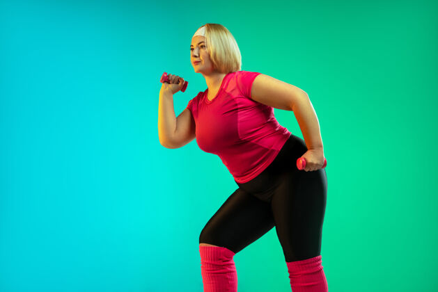 拉伸年轻的白人大号女模特在霓虹灯下的渐变绿色背景下训练用重量做锻炼运动的概念 健康的生活方式 积极的身体 平等健康专业人