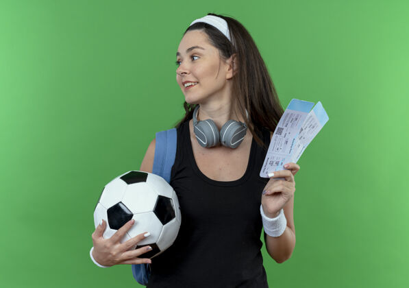 微笑戴着头带 带着耳机 背着背包 拿着足球和机票 微笑着站在绿墙上的年轻健身女士空气头带健身