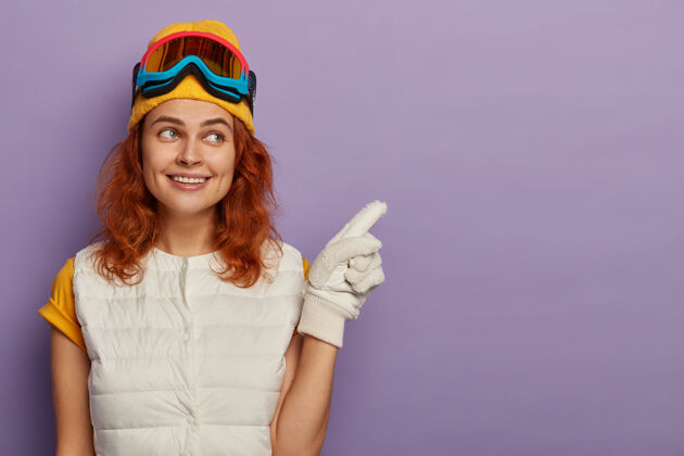 空白迷人的红发女人戴着暖和的帽子 滑雪镜和白色套头衫滑雪者指点青少年