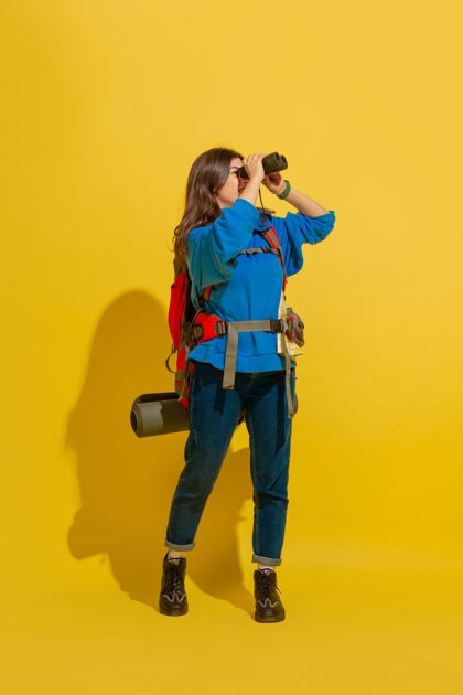 人黄色摄影棚墙上 一个带着包和望远镜的快乐的年轻旅游女孩的画像年轻人方式快乐